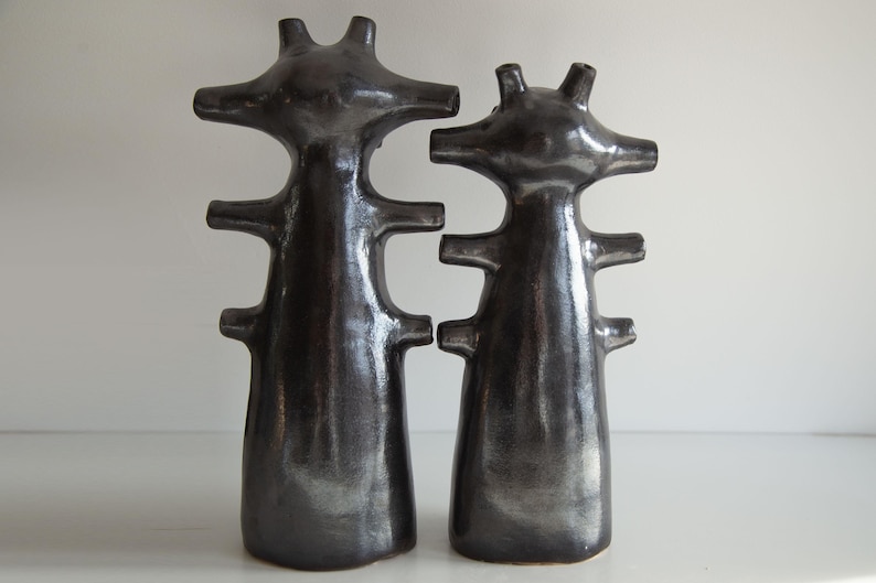 Op de afbeelding: Twee donkergrijze keramische sculpturen met een abstract ontwerp. Elke sculptuur heeft een slank lichaam met horizontale uitsteeksels en een kopachtig bovenstuk met drie punten. De sculpturen staan op een wit oppervlak.