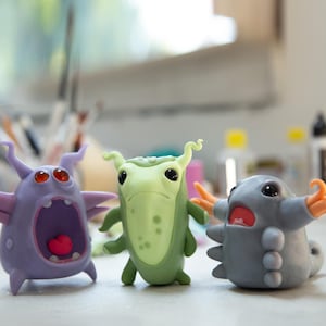 May include: Three colorful, cartoonish monster figurines. The purple monster has open mouth with a pink heart, the green monster has a serious expression, and the gray monster has orange tentacles. The figurines are on a white surface.