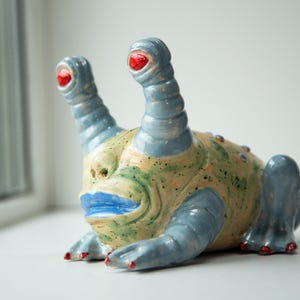 May include: A ceramic sculpture of a fantastical creature with blue, green, and tan hues. It has two blue, ribbed antennae with red eyes, a blue mouth, and blue feet with red accents. The creature is resting on a white surface.
