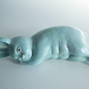 Op de afbeelding: Een lichtblauw keramisch beeldje van een konijn dat op zijn zij ligt. Het konijn heeft lange oren, een gladde afwerking en een mensachtig bovenlichaam met gedefinieerde buikspieren. Het figuur rust met het hoofd op de voorpoten.