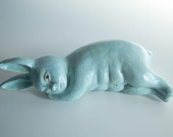 Könnte beinhalten: Eine hellblaue Keramikskulptur eines auf der Seite liegenden Hasen. Der Hase hat lange Ohren, eine glatte Oberfläche und einen menschenähnlichen Oberkörper mit definierten Bauchmuskeln. Die Figur ruht ihren Kopf auf ihren Vorderpfoten.