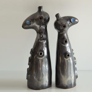 Op de afbeelding: Twee abstracte keramische sculpturen met een donkere, metallic afwerking. Elk heeft een unieke, langwerpige vorm met meerdere cirkelvormige openingen en een klein, blauw accent. De sculpturen worden op een wit oppervlak weergegeven.