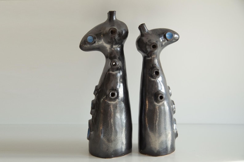 Op de afbeelding: Twee abstracte keramische sculpturen met een donkere, metallic afwerking. Elk heeft een unieke, langwerpige vorm met meerdere cirkelvormige openingen en een klein, blauw accent. De sculpturen worden op een wit oppervlak weergegeven.