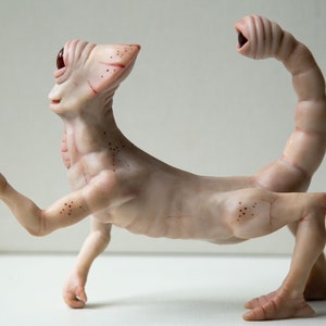 May include: A hand-crafted, pale pink, humanoid sculpture with a single large eye and a long, segmented tail. The sculpture has a detailed, textured surface and appears to be made of polymer clay.