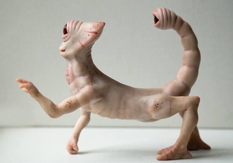 May include: A hand-crafted, pale pink, humanoid sculpture with a single large eye and a long, segmented tail. The sculpture has a detailed, textured surface and appears to be made of polymer clay.
