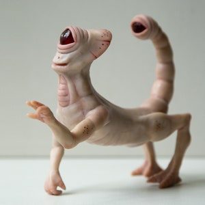 May include: A white, humanoid sculpture with a single red eye, a long, segmented tail, and four legs. The sculpture is made of a smooth, fleshy material and has a slightly unsettling appearance.