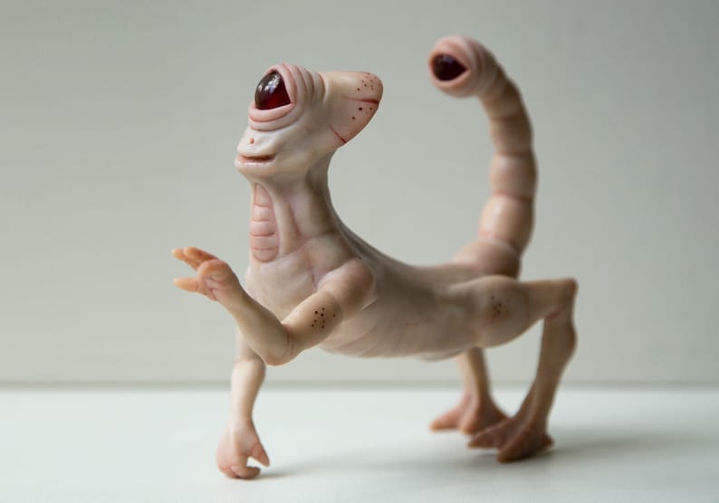 May include: A white, humanoid sculpture with a single red eye, a long, segmented tail, and four legs. The sculpture is made of a smooth, fleshy material and has a slightly unsettling appearance.