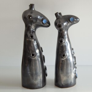 Op de afbeelding: Twee abstracte keramische sculpturen met een donkere, metallic afwerking. Elk heeft een unieke, langwerpige vorm met cirkelvormige openingen en blauwe accenten. De sculpturen worden op een wit oppervlak weergegeven.