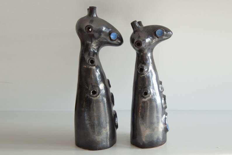 Op de afbeelding: Twee abstracte keramische sculpturen met een donkere, metallic afwerking. Elk heeft een unieke, langwerpige vorm met cirkelvormige openingen en blauwe accenten. De sculpturen worden op een wit oppervlak weergegeven.