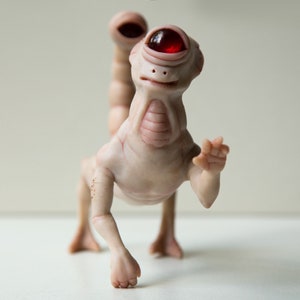 May include: A small, white, humanoid sculpture with a single red eye and a tail. The sculpture is posed in a walking position with one arm raised in a wave.