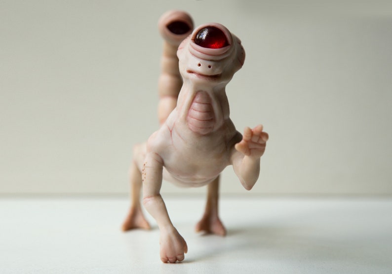 May include: A small, white, humanoid sculpture with a single red eye and a tail. The sculpture is posed in a walking position with one arm raised in a wave.