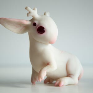 Könnte beinhalten: Eine weiße Figur einer Fabelwesen mit großen, dunkelroten Augen und kleinen Geweihen. Die Kreatur hat große Ohren mit rosa Adern und rosa Akzenten an Pfoten und Schnauze. Sie sitzt.