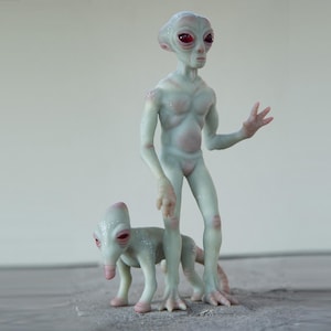 K&ouml;nnte beinhalten: Eine graue Alien-Figur mit roten Augen und eine kleine graue Alien-Haustierfigur. Beide Figuren haben eine strukturierte Oberfl&auml;che.