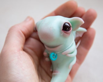 Könnte beinhalten: Eine kleine, hellblaue Drachenfigur mit großen, dunklen Augen und einer türkisfarbenen Spiralenkette. Der Drache hat ein zartes, fantasievolles Aussehen.
