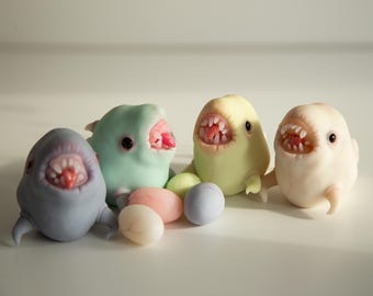 Könnte beinhalten: Vier skurrile Monsterfiguren in Pastelltönen von Blau, Grün, Gelb und Weiß, jede mit offenem Mund, der Zähne und Zungen zeigt. Kleine, runde Augen und pastellfarbene Eier vervollständigen die Szene.