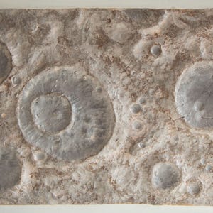 Könnte beinhalten: Ein rechteckiges, strukturiertes Kunstwerk mit einem Mondoberflächendesign. Das Stück zeigt Krater unterschiedlicher Größe in Grau- und Brauntönen mit einer rauen, unebenen Oberfläche. Das Kunstwerk erinnert an die Mondlandschaft.