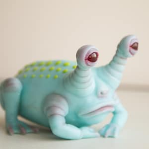 May include: A light blue, alien-like figurine with large, bulbous eyes and a grumpy expression. The figurine has two large, upward-pointing eyes with red pupils. The body is a light blue with yellow spots on the back.
