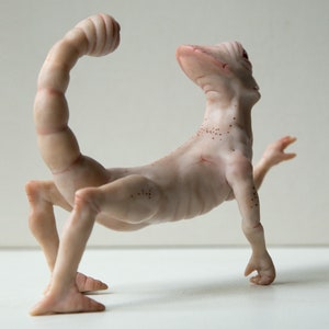 May include: A nude-colored ceramic sculpture of a scorpion-like creature with a long, segmented tail and four legs. The creature has a human-like head and torso, and its body is covered in small, brown spots.