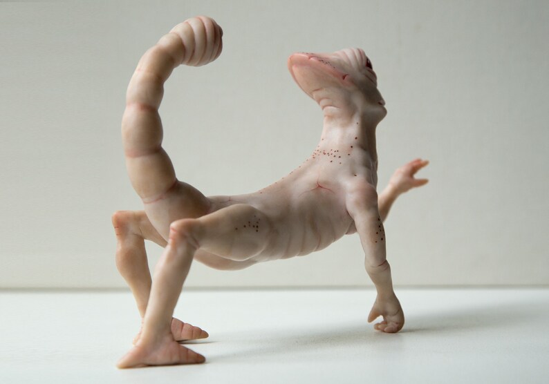 May include: A nude-colored ceramic sculpture of a scorpion-like creature with a long, segmented tail and four legs. The creature has a human-like head and torso, and its body is covered in small, brown spots.