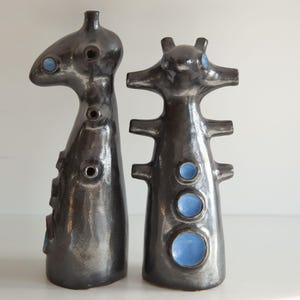 Op de afbeelding: Twee abstracte keramische sculpturen. De sculpturen zijn donkergrijs met een glanzende afwerking en hebben blauwe cirkelvormige accenten. De vormen zijn hoog en slank, met unieke vormen en openingen, wat een moderne kunstesthetiek suggereert.