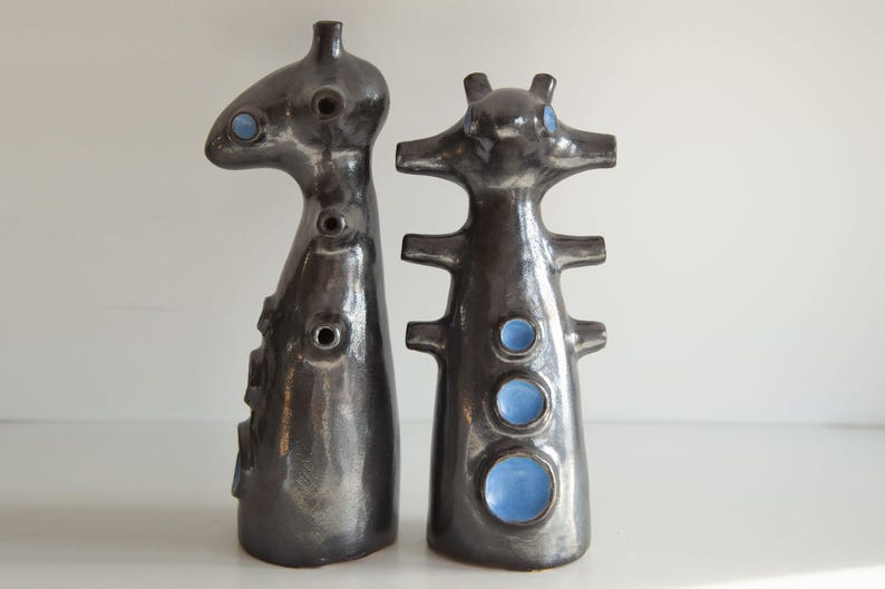 Op de afbeelding: Twee abstracte keramische sculpturen. De sculpturen zijn donkergrijs met een glanzende afwerking en hebben blauwe cirkelvormige accenten. De vormen zijn hoog en slank, met unieke vormen en openingen, wat een moderne kunstesthetiek suggereert.
