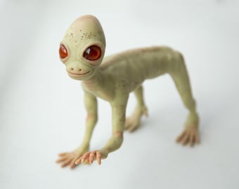 以下が含まれることがあります： 赤い目をした緑色のヒューマノイドの置物。歯を見せて笑っている。置物は細身の体と長く細い手足を持ち、歩いているポーズをとっている。頭は少し横に傾いている。