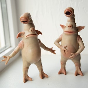 May include: Two whimsical, light-skinned alien figurines with elongated heads and expressive faces. The figures have small ears, arms, and feet, and are posed in different stances. The figurines are displayed on a white surface near a window.