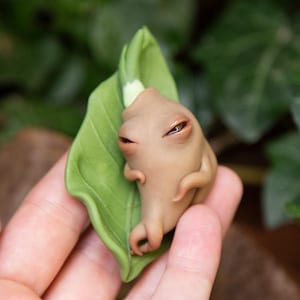 May include: A tan-colored, stylized figurine with closed eyes and small arms, cradled by a large, green leaf. The figurine has a smooth texture and is set against a blurred green background.