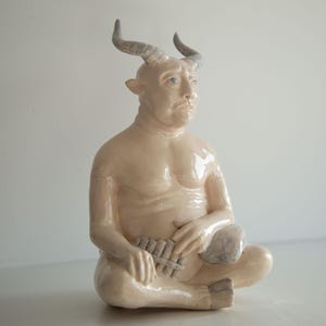 Puede incluir: Una escultura de cerámica de una figura sentada con rostro humano, cuernos y patas de cabra. La figura es de color beige claro con cuernos grises y sostiene una flauta de pan. La escultura está en una pose meditativa.