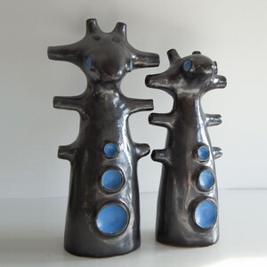 Op de afbeelding: Twee donkergrijze keramische sculpturen met een glanzende afwerking. Elk heeft een unieke, abstracte vorm met meerdere uitsteeksels en cirkelvormige blauwe accenten. De sculpturen zijn verschillend van grootte en staan op een wit oppervlak.