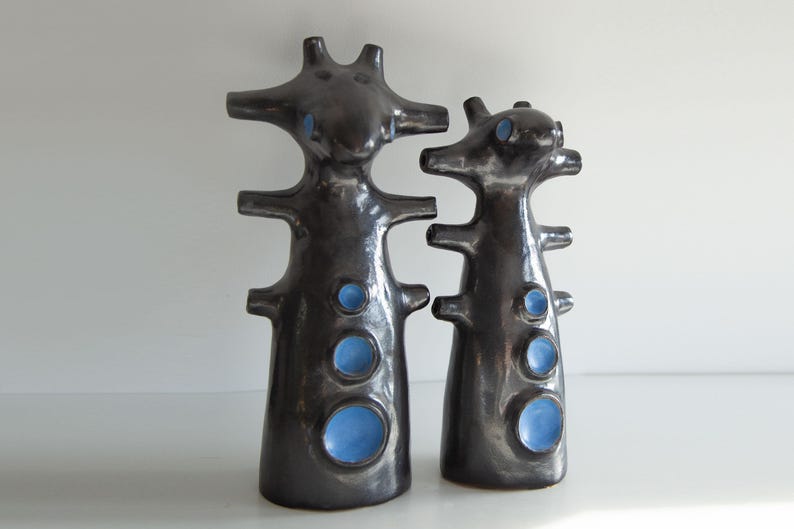 Op de afbeelding: Twee donkergrijze keramische sculpturen met een glanzende afwerking. Elk heeft een unieke, abstracte vorm met meerdere uitsteeksels en cirkelvormige blauwe accenten. De sculpturen zijn verschillend van grootte en staan op een wit oppervlak.