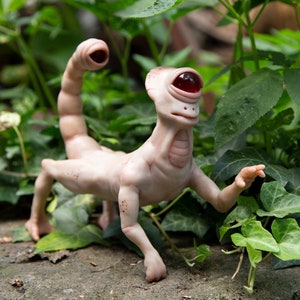 May include: A hand-crafted, beige-colored, humanoid sculpture with a single red eye and a scorpion tail. The sculpture is posed in a walking position with one arm raised. The sculpture is set against a backdrop of green foliage.