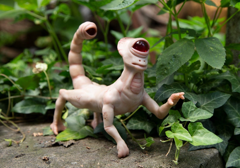 May include: A hand-crafted, beige-colored, humanoid sculpture with a single red eye and a scorpion tail. The sculpture is posed in a walking position with one arm raised. The sculpture is set against a backdrop of green foliage.