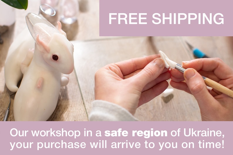 May include: A white ceramic dragon sculpture with a glass wing. The dragon is being painted with a brush by a person's hand. The text "FREE SHIPPING" is displayed in a pink banner above the image. The text "Our workshop in a safe region of Ukraine, your purchase will arrive to you on time!" is displayed in a pink banner below the image.
