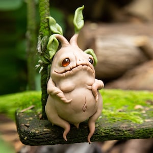 Può includere: Una piccola scultura in argilla beige di una creatura con un corpo rotondo, quattro zampe tozze e una piccola testa con foglie. La creatura ha grandi occhi arancioni e una piccola bocca marrone. È seduta su una superficie di legno ricoperta di muschio.