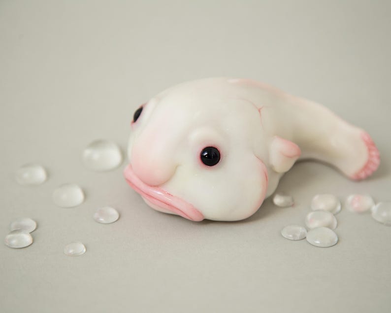 May include: A white, hand-painted sculpture of a blobfish with black eyes and a pink mouth. The sculpture is lying on its side on a light gray surface.