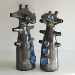 Op de afbeelding: Twee abstracte keramische sculpturen met een donkere, metallic afwerking. Elk heeft een unieke, organische vorm met meerdere uitsteeksels en openingen. De sculpturen hebben blauwe accenten en worden gepresenteerd tegen een witte achtergrond.