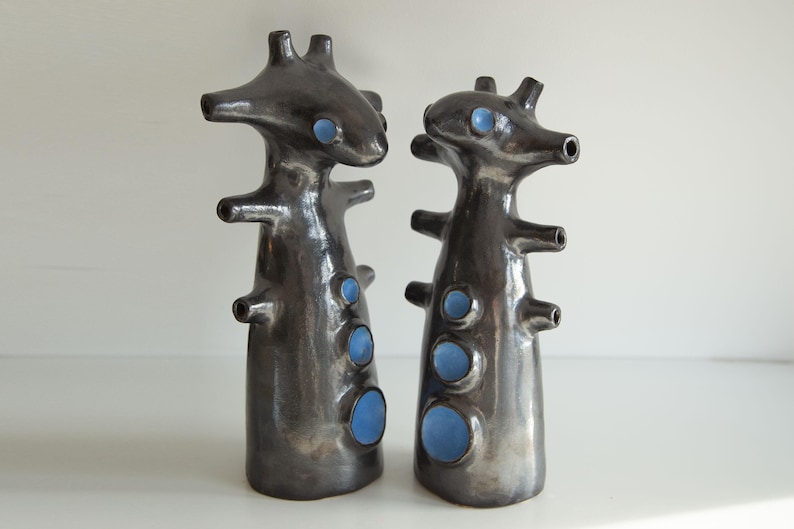 Op de afbeelding: Twee abstracte keramische sculpturen met een donkere, metallic afwerking. Elk heeft een unieke, organische vorm met meerdere uitsteeksels en openingen. De sculpturen hebben blauwe accenten en worden gepresenteerd tegen een witte achtergrond.