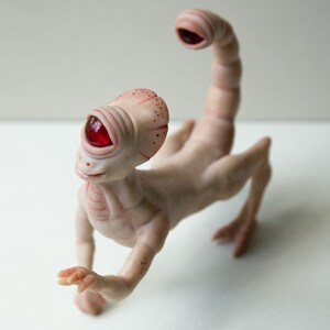 May include: A small, pale pink, humanoid sculpture with a single red eye and a scorpion tail. The sculpture is made of clay and has a whimsical, otherworldly appearance.