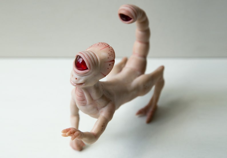 May include: A small, pale pink, humanoid sculpture with a single red eye and a scorpion tail. The sculpture is made of clay and has a whimsical, otherworldly appearance.
