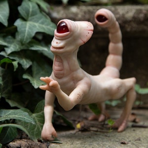 May include: A pale, humanoid sculpture with a single large red eye and a scorpion tail. The sculpture is made of a smooth, white material and has a slightly whimsical expression.