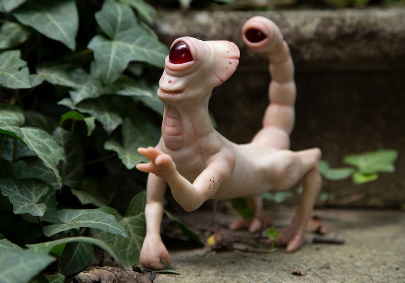 May include: A pale, humanoid sculpture with a single large red eye and a scorpion tail. The sculpture is made of a smooth, white material and has a slightly whimsical expression.