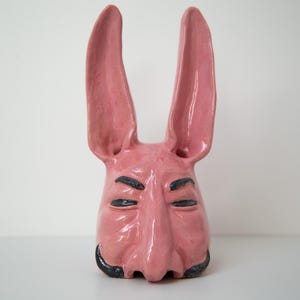 May include: A pink ceramic rabbit mask with black painted eyes and a moustache. The mask has a long, pointed nose and large ears.
