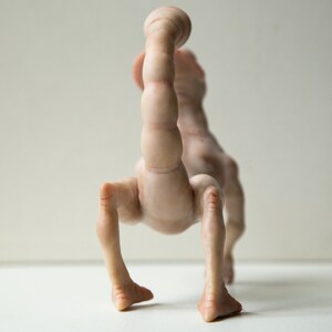 May include: A nude-colored ceramic sculpture of a creature with a long, segmented tail and four legs. The creature is standing on a white surface.
