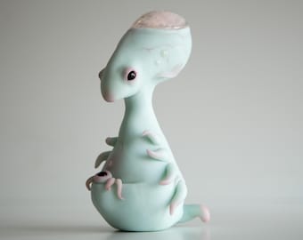 May include: A quirky, pale-blue alien figurine with pink detailing. The figure features large eyes, a rounded head, and several tentacles. The sculpture seems to be crafted from ceramic or a comparable material, with a smooth, glossy surface.