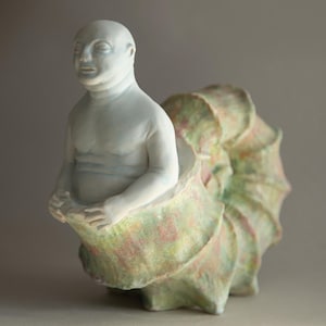 May include: A ceramic sculpture of a bald figure emerging from a large, shell-like form. The figure is pale with subtle blue shading, and the shell is a mix of green, pink, and yellow hues. The sculpture has a unique, artistic design.
