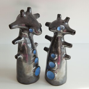Op de afbeelding: Twee abstracte keramische sculpturen met een donkere metallic afwerking en blauwe accenten. Elk stuk heeft een unieke, organische vorm met meerdere uitsteeksels en openingen, wat een moderne, artistieke esthetiek cre&euml;ert.