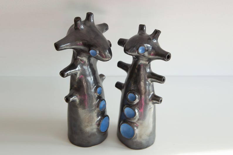 Op de afbeelding: Twee abstracte keramische sculpturen met een donkere metallic afwerking en blauwe accenten. Elk stuk heeft een unieke, organische vorm met meerdere uitsteeksels en openingen, wat een moderne, artistieke esthetiek cre&euml;ert.