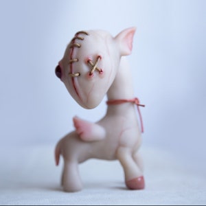 May include: A small, white, winged creature with stitched-up eyes and a red ribbon around its neck. The creature is made of a soft, pliable material and has a whimsical, slightly unsettling appearance.