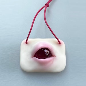 Op de afbeelding: Een witte keramische hanger in de vorm van een mond met een rood en roze interieur en een rode edelsteen in het midden. De hanger is bevestigd aan een rood koord.
